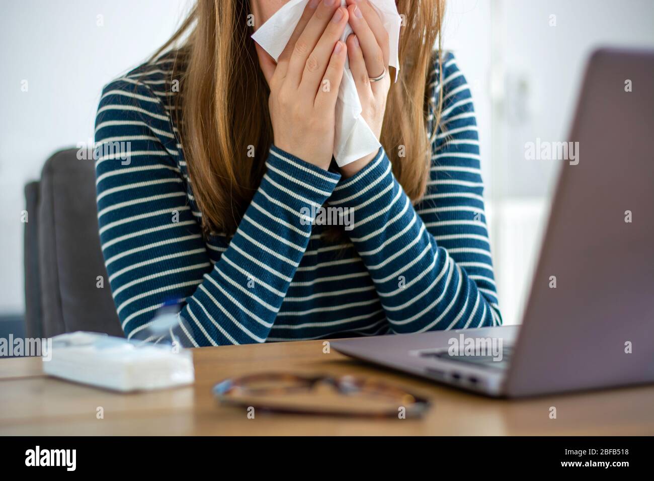 Sick woman working on laptop at home, covering nose and mouth when coughing and sneezing in paper tissue, ill freelance woman with cold working Stock Photo