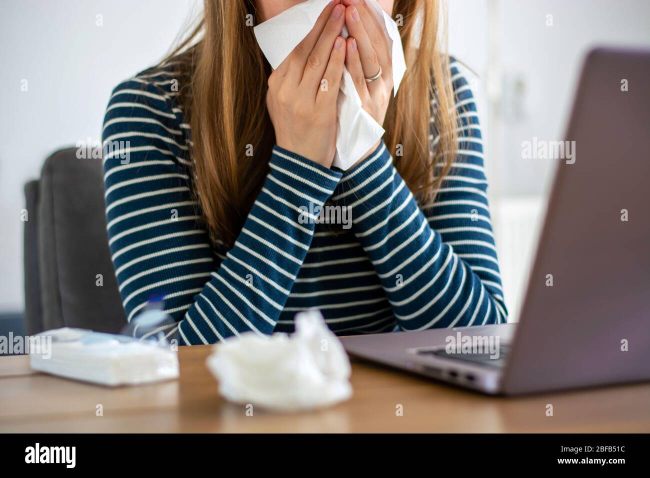 Sick woman working on laptop at home, covering nose and mouth when coughing and sneezing in paper tissue, ill freelance woman with cold working Stock Photo