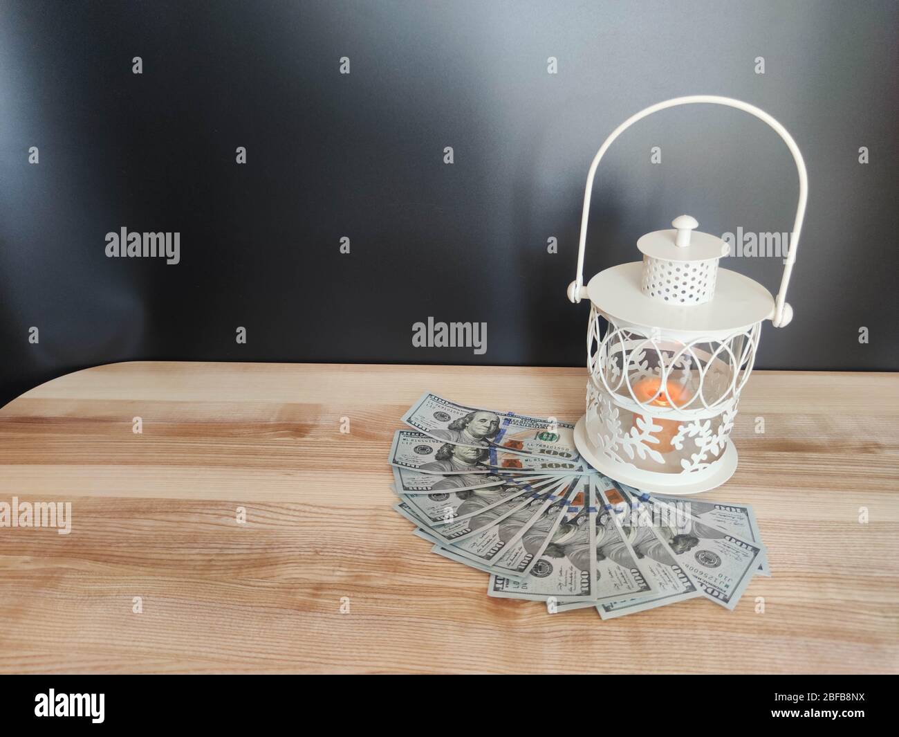 White lantern with an orange candle, 100 dollar banknotes and a mask on the table with black backdrop. Concept - Ramadan kareem holiday celebration in Stock Photo