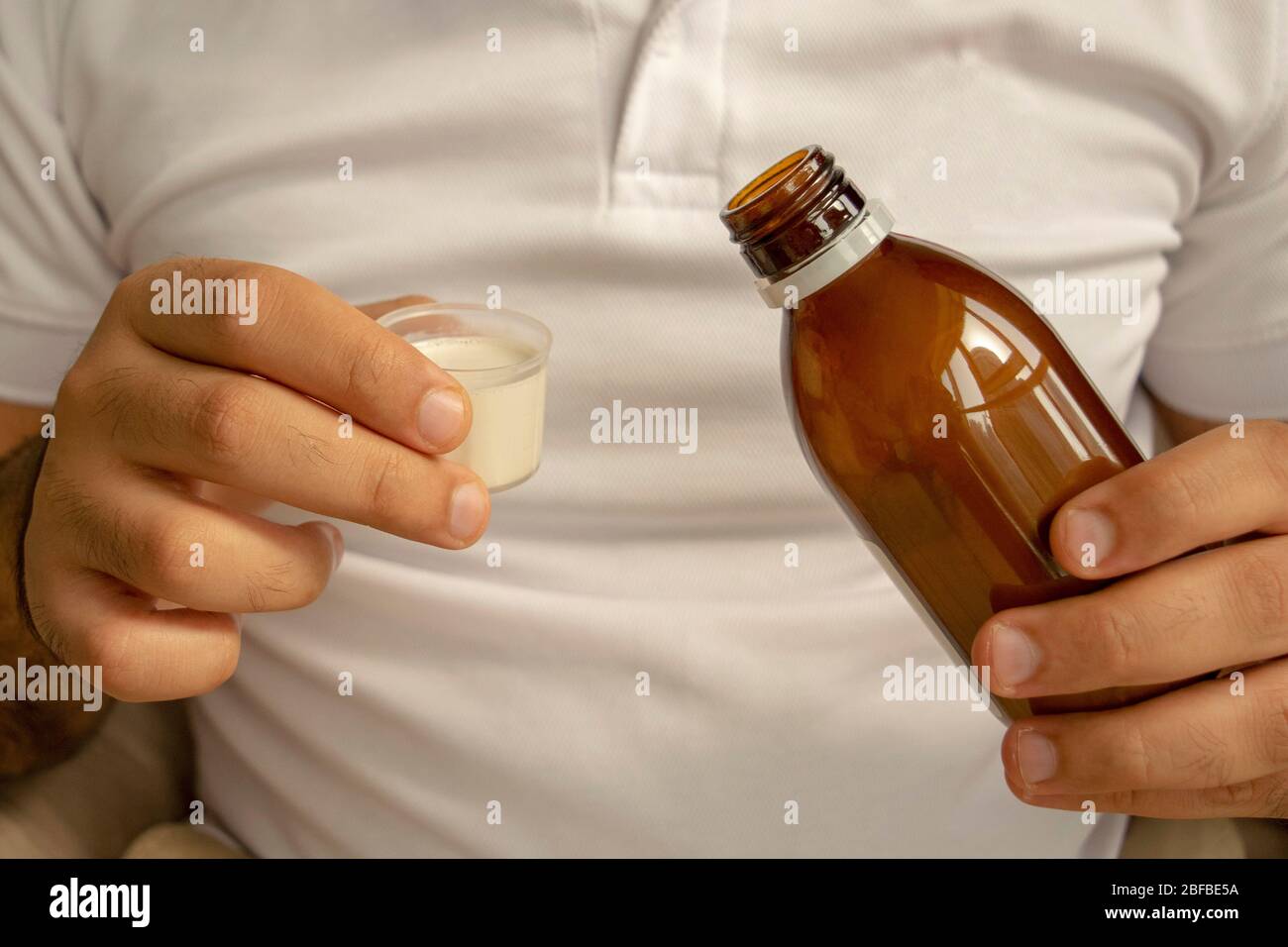 Man in a white t-shirt is pouring medical syrup in a measuring cup. A young man holds a bottle of syrup with a cup. Diarrhea, colitis, dysentery drug.  oral s Stock Photo