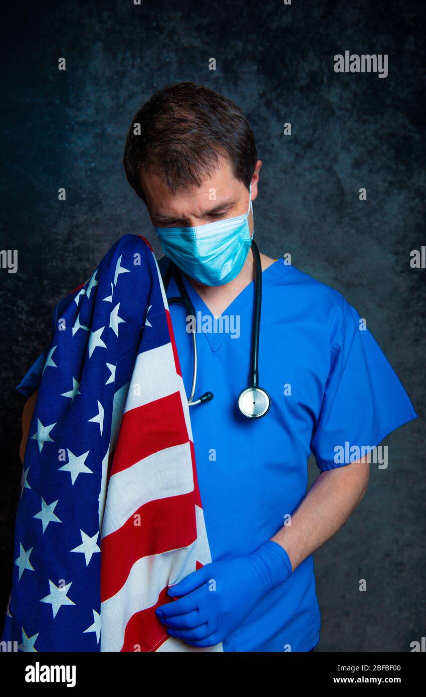 Sad/pensive, male doctor in blue hospital scrubs with face mask and stethoscope, holding the Stars & Stripes American flag close to his chest. Stock Photo
