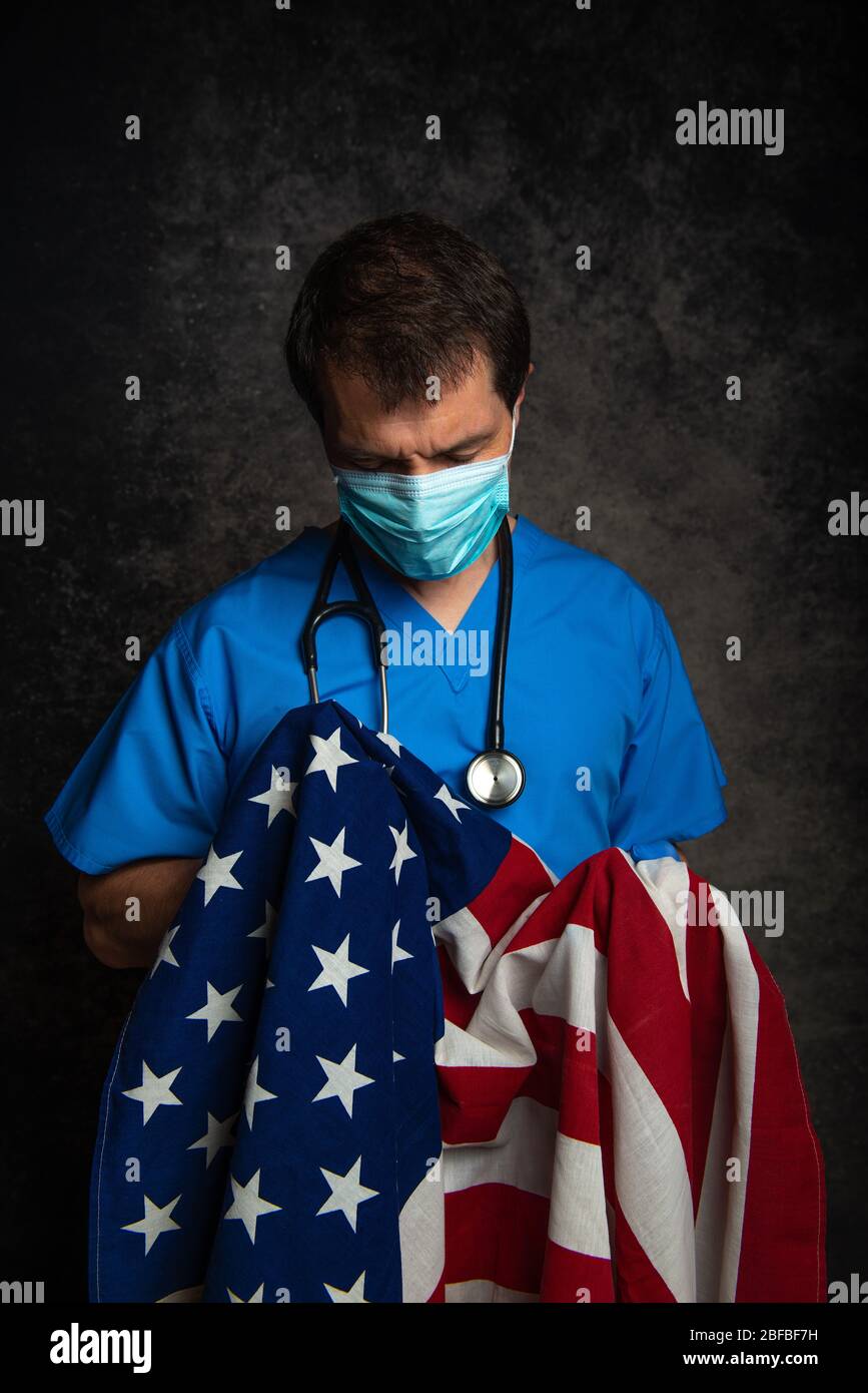 Sad/pensive, male doctor in blue hospital scrubs with face mask and stethoscope, holding the Stars & Stripes American flag close to his chest. Stock Photo