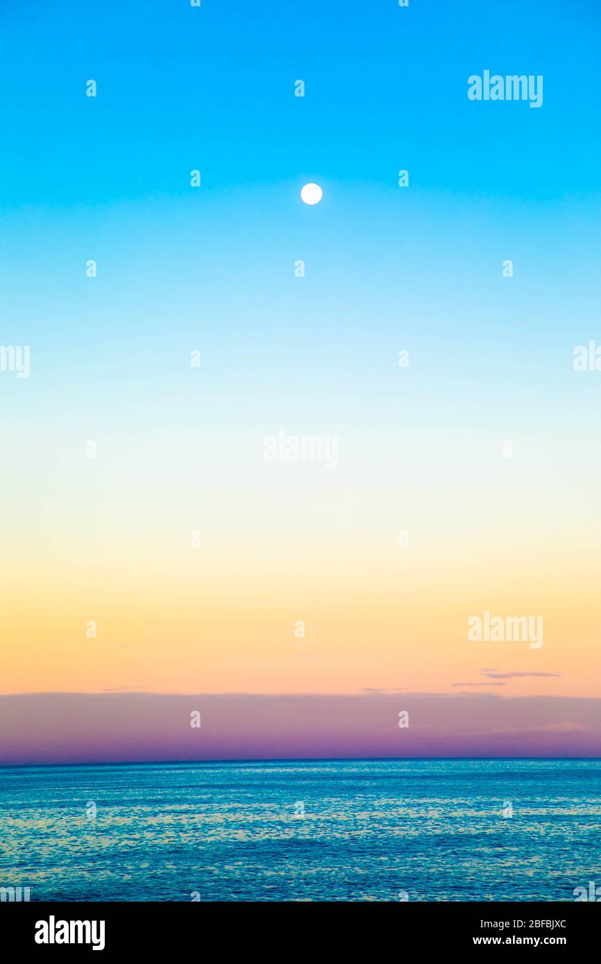 Full moon setting over the ocean during sunrise with pastel colors Stock Photo