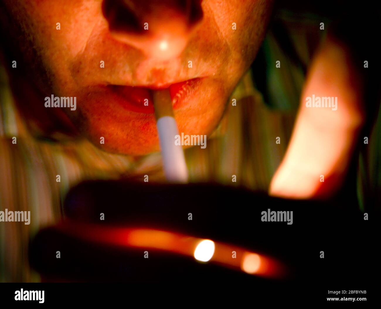 A close-up picture of a man lighting a cigarette, his face solely lit by the light of a lighter Stock Photo