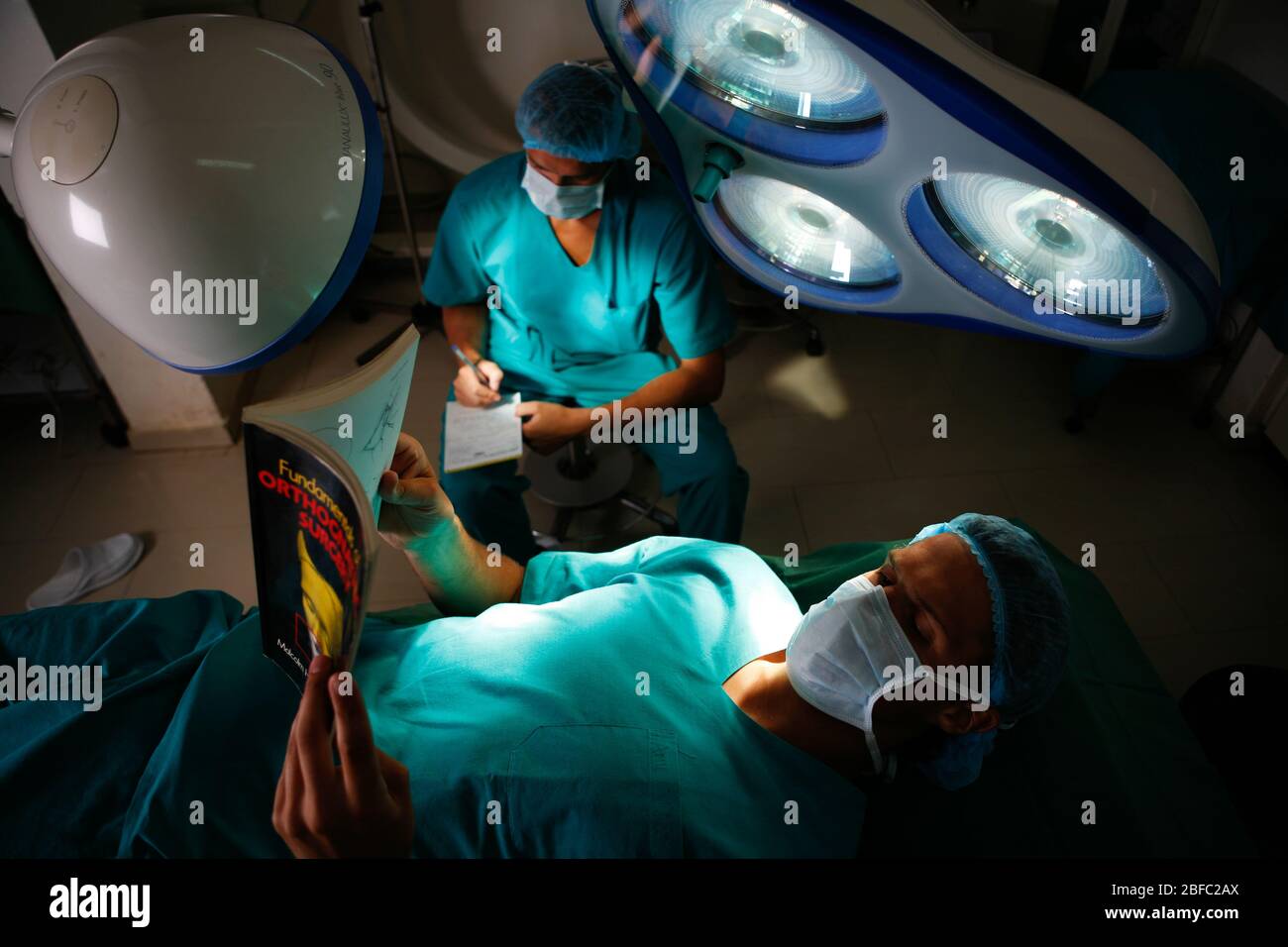A surgeon reading a book whilst laying on an operating table. A second surgeon can be seen writing notes in the background. Stock Photo