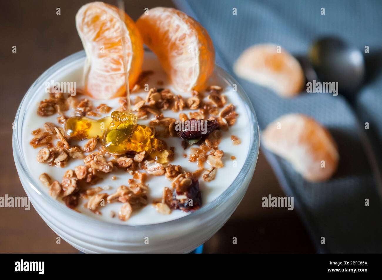 Close up image of a cup of sugarfree yogurt with two pieces of tangerine granola and honey bee, with blurry tangerines and a spoon on the background Stock Photo