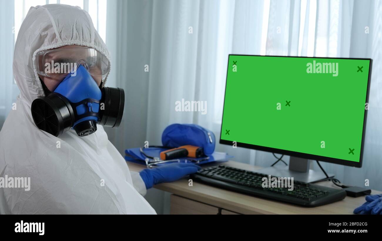 Medium shot. View to the display. Medical Doctor In Hazard Suit Looking to Camera Sitting Next to Blank Monitor. Green Screen Mock-up Display. Profess Stock Photo