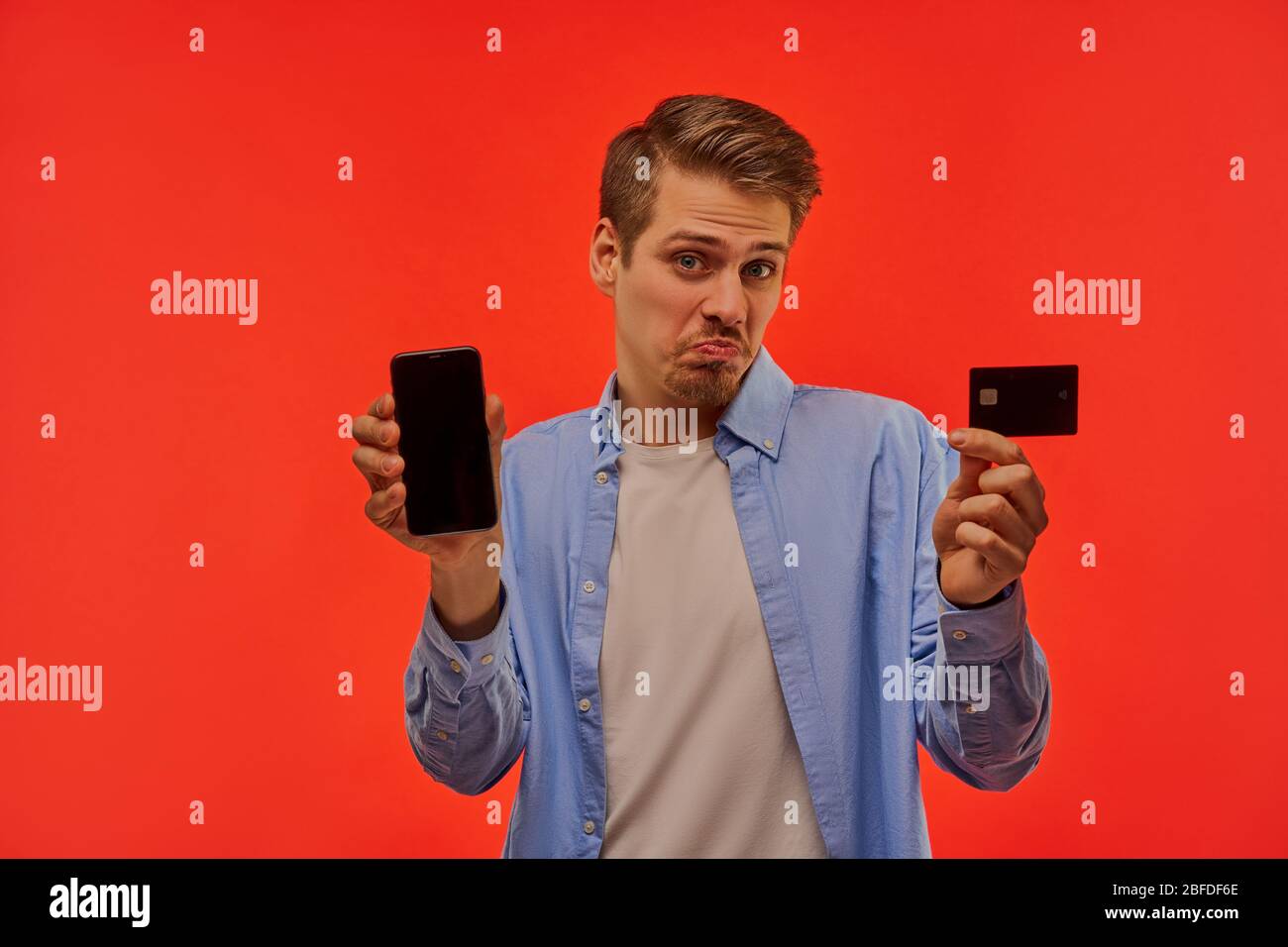 Disappointed guy with a beard shows a smartphone and a credit card. Stock Photo