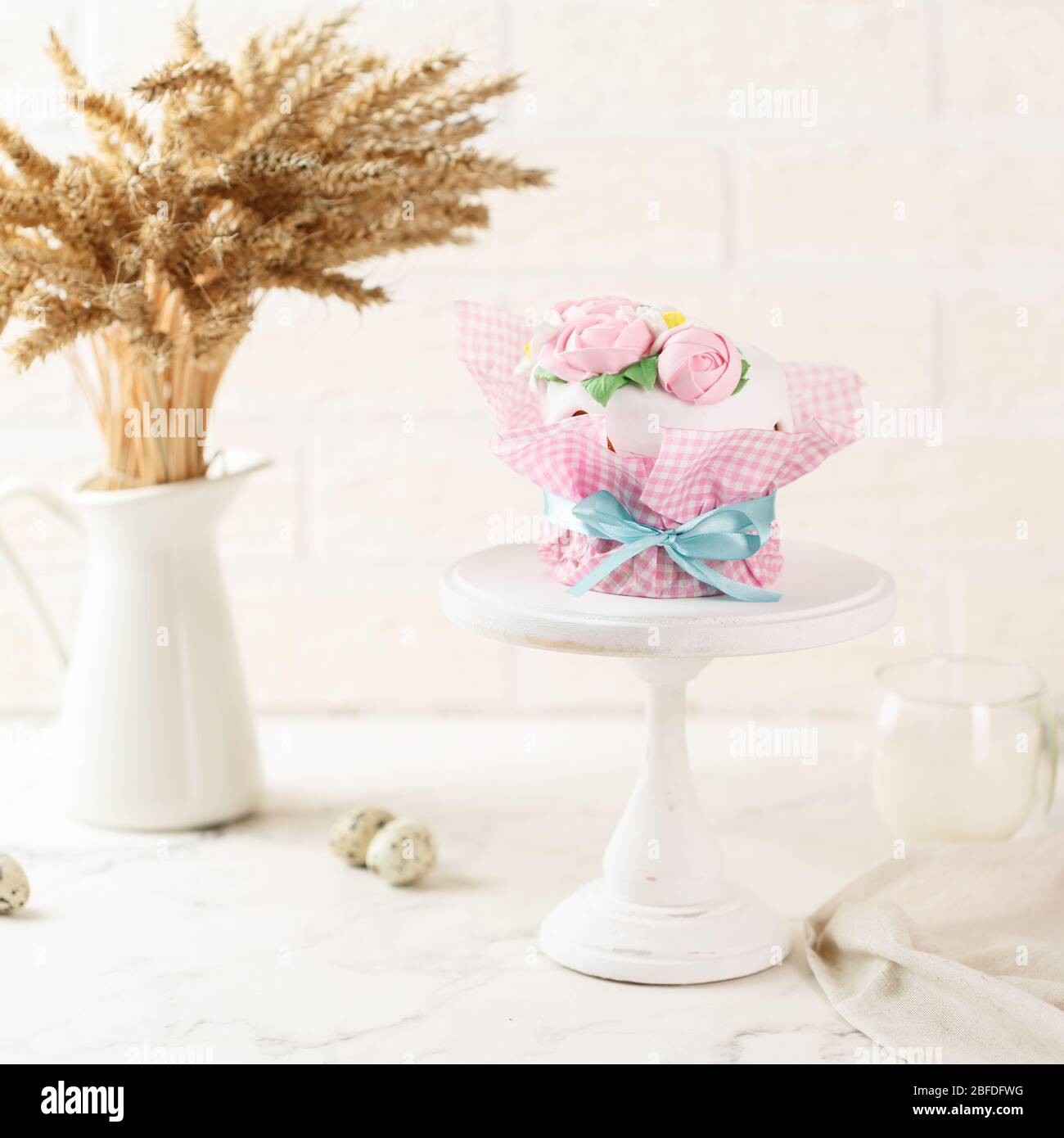 Easter cake in pink paper packaging with blue ribbon on the grey plate. Homemade festive pie with frosting and mastic floral decoration. Bunch of whea Stock Photo