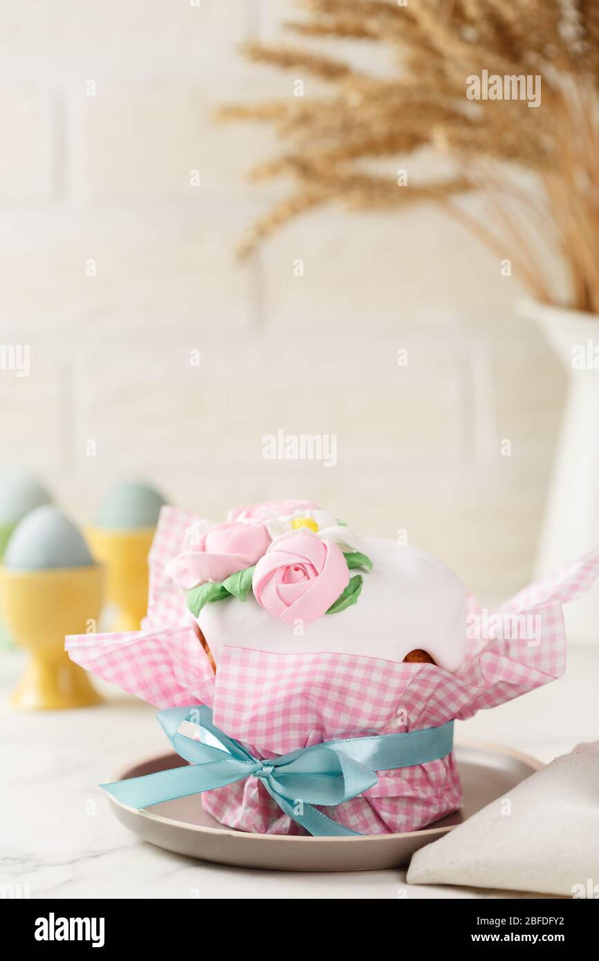 Easter cake in pink paper packaging with blue ribbon on the grey plate. Homemade festive pie with frosting and mastic floral decoration. Bunch of whea Stock Photo