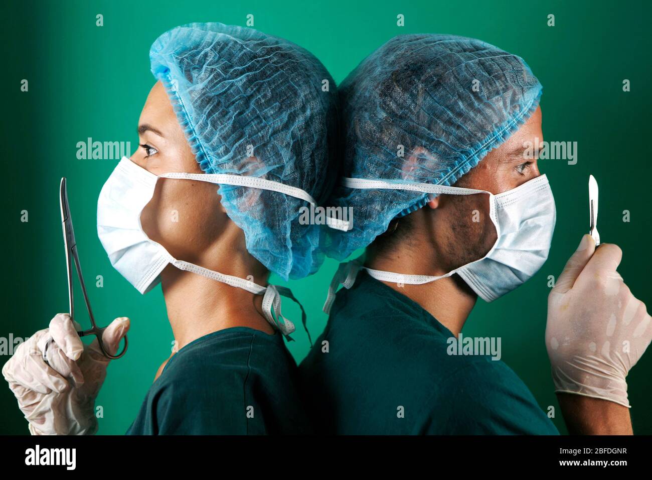 Profle of two hospital surgeons standing back to back. The female surgeon on the left holds a pair of forceps in front of her face, the male surgeon o Stock Photo