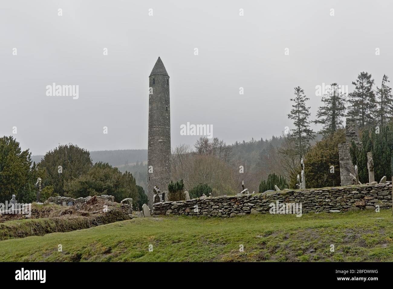Spooky old cemetery with round tower in the fog in Glendalough monastic site with stone wall and mountain forest in the background, Dublin mountains,W Stock Photo