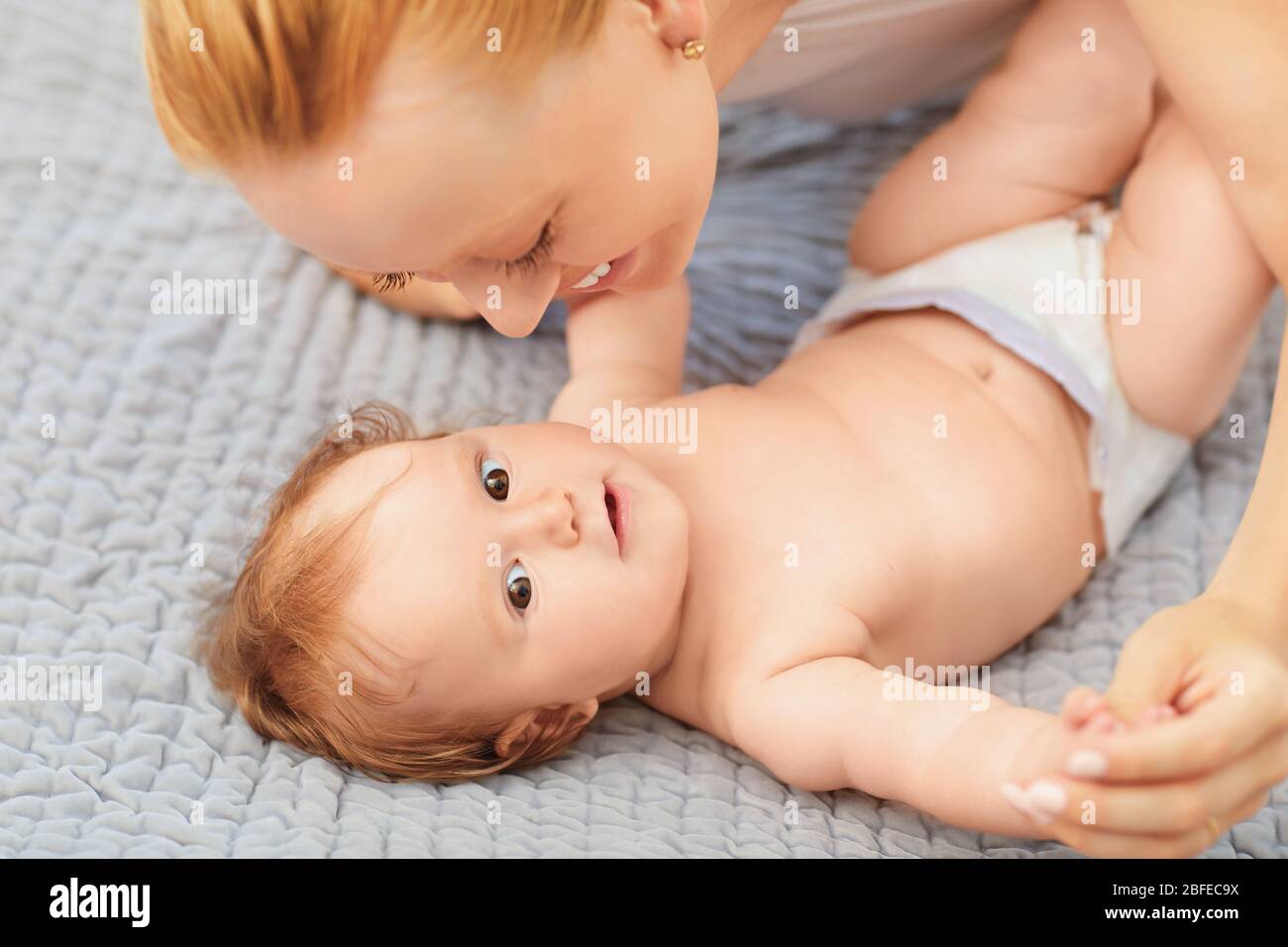 Happy mother smiles at baby lying on bed in room. Stock Photo