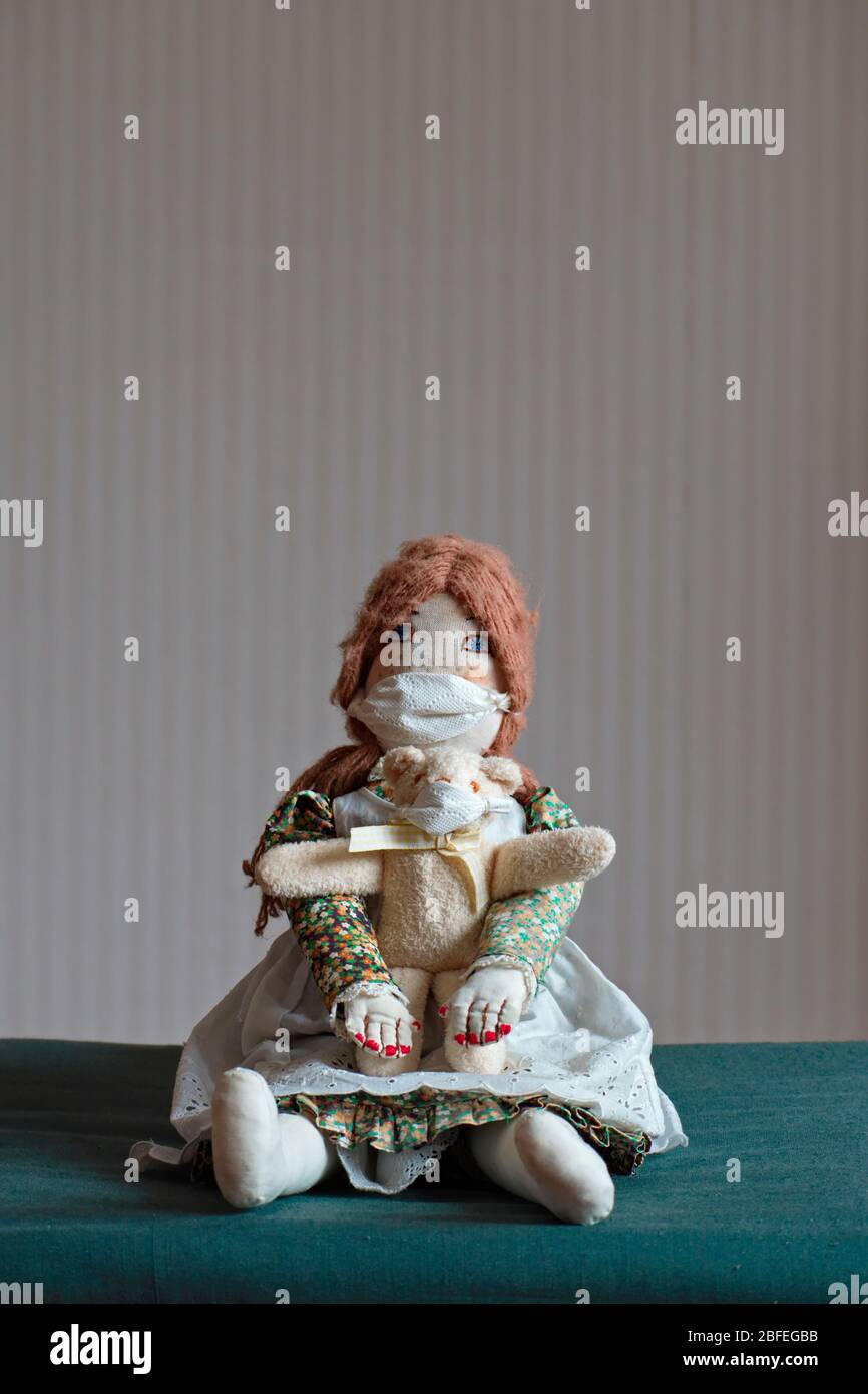 A handmade old-fashioned female doll clothed in a dress with a pinafore is sitting on a chair, wearing a protective mask. She holds a small teddy bear Stock Photo