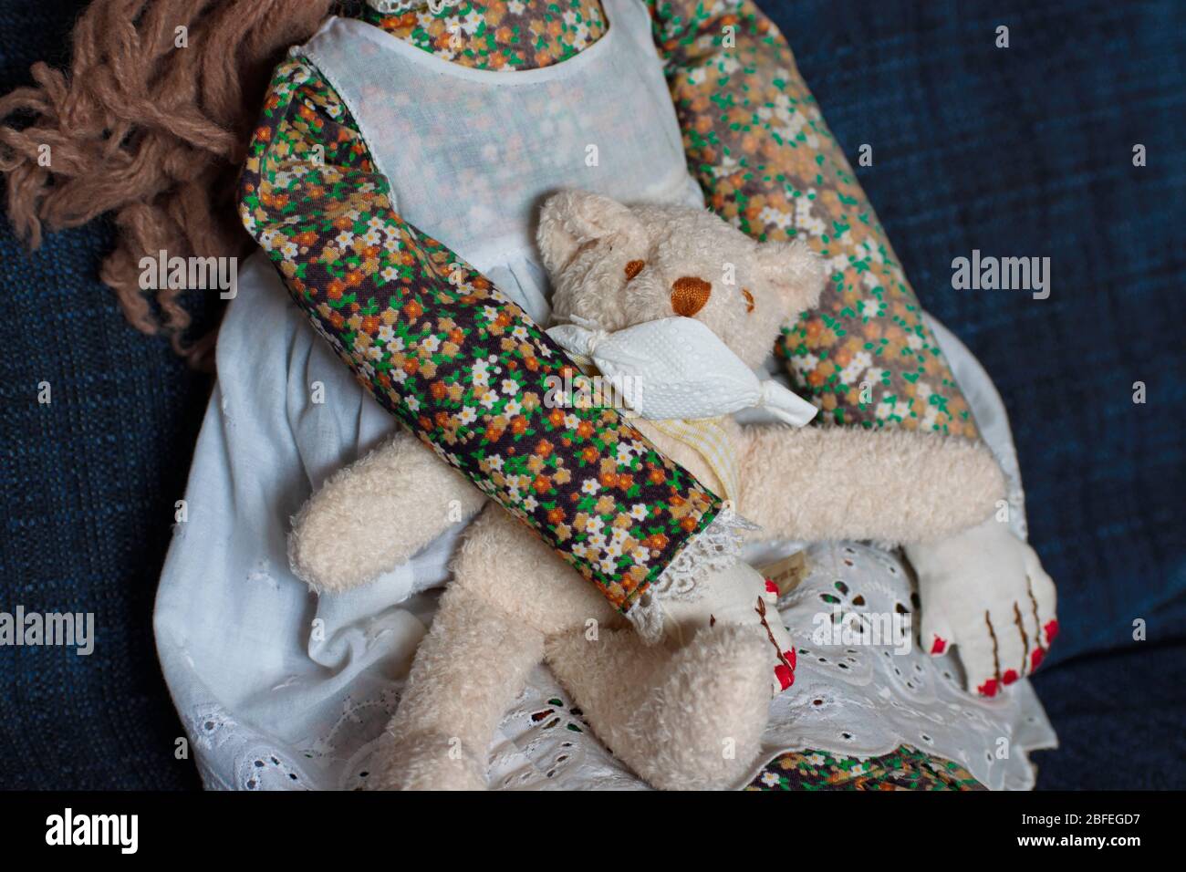 A handmade old-fashioned female doll clothed in a dress with a pinafore is sitting on a blue sofa.She holds a small teddy bear in her arms, he's  wear Stock Photo