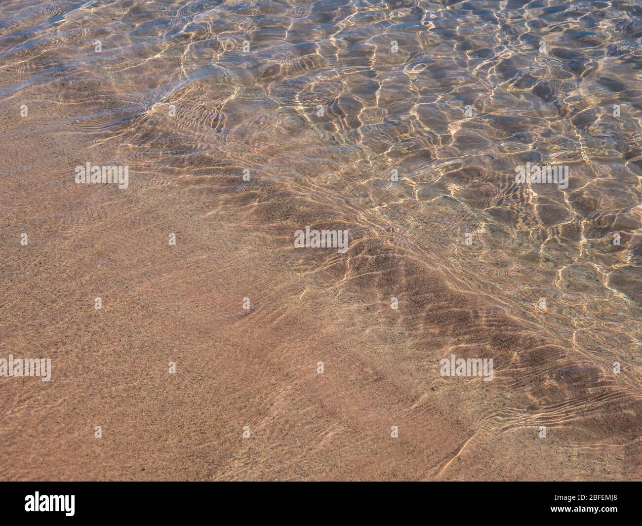 Horizontal detail of a beach with smooth waves of sparkling water moving in diagonal lines over fine sand. Stock Photo