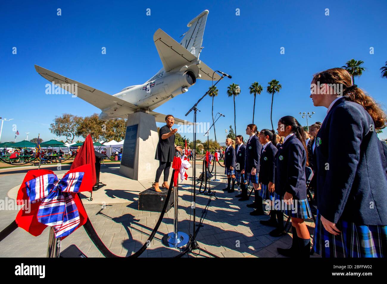 A Catholic school girls' choir is directed by their African American leader as they sing patriotic songs at Veterans Day observances in Costa Mesa, CA. The A4M Skyhawk aircraft is a patriotic monument. Stock Photo