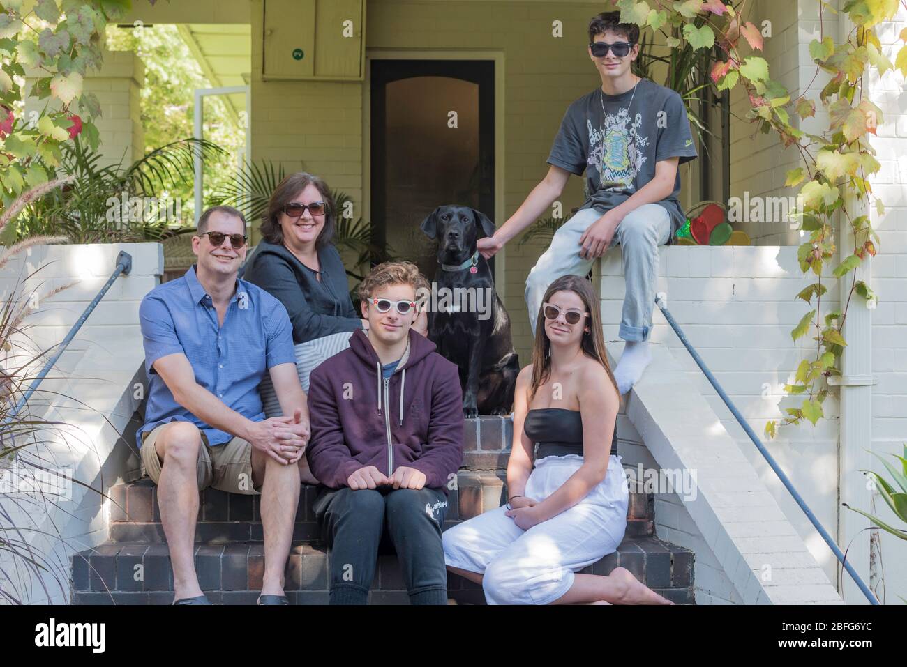 A smiling Australian family of husband wife daughter and two sons plus pet dog all wearing sunglasses, sit at the front steps of their Sydney home. Stock Photo