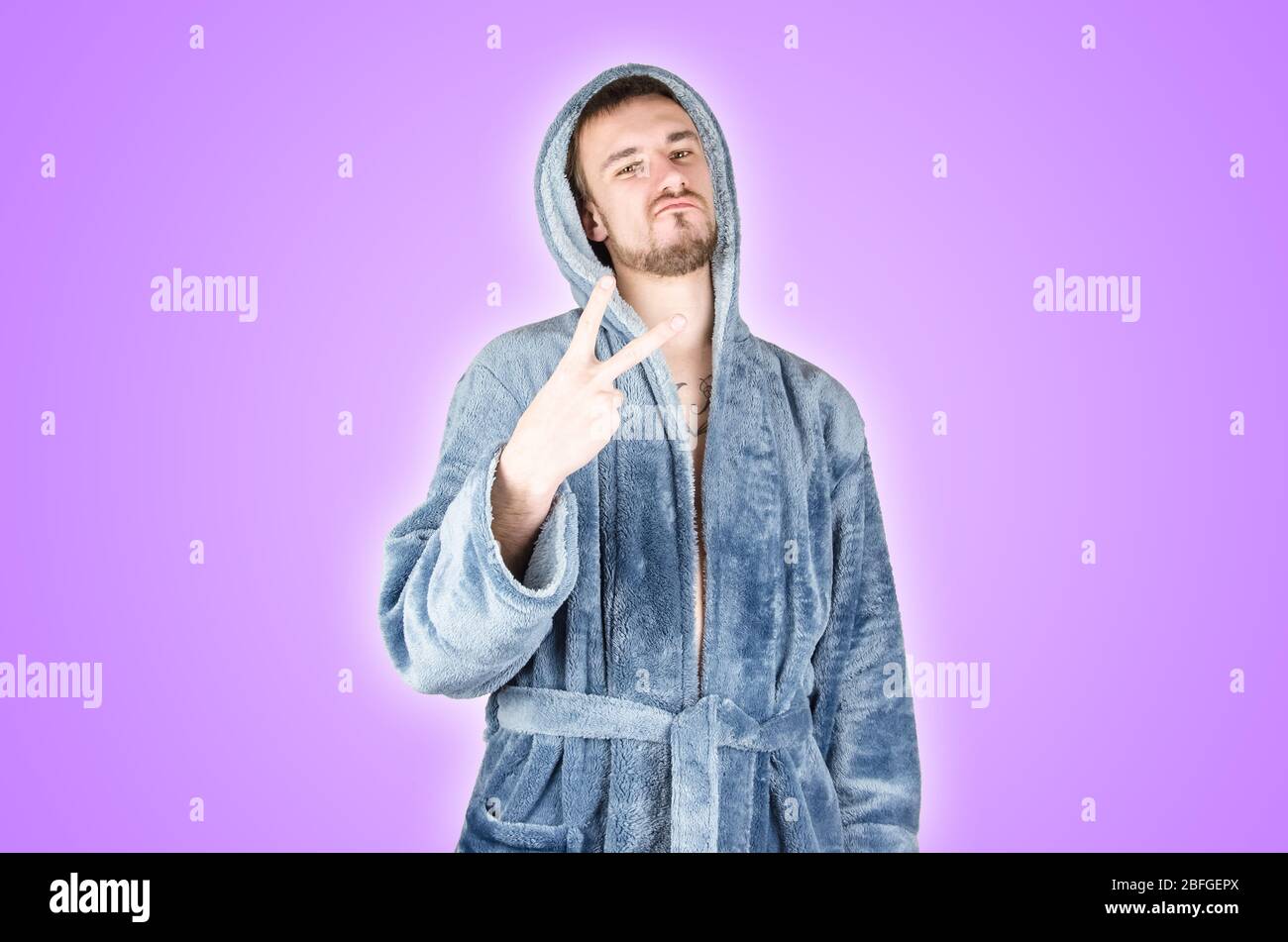 Portrait of young caucasian bearded man in blue bathrobe shows victory or peace gesture isolated on purple background. Happy emotion Stock Photo