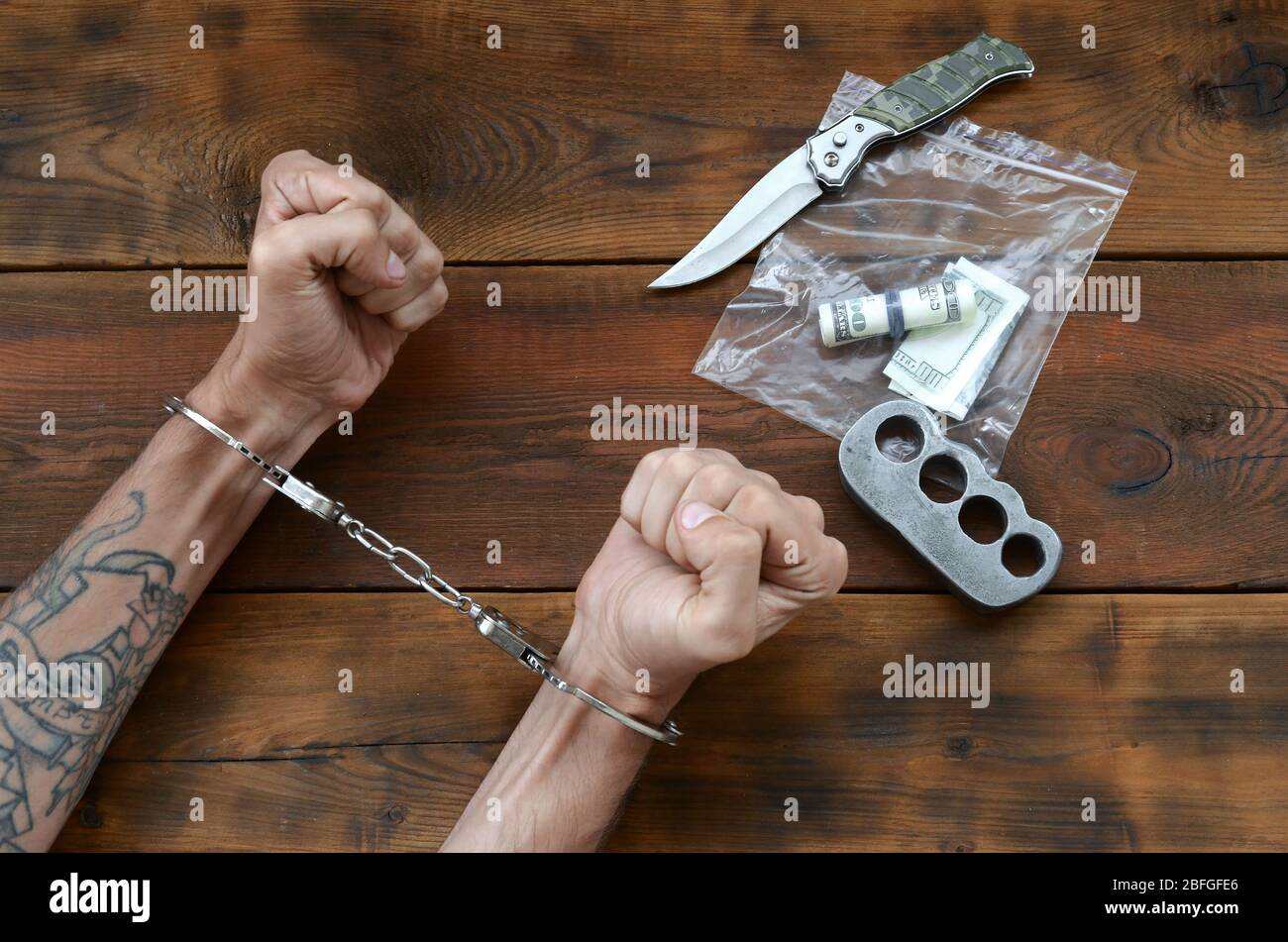 Cuffed hands of tattooed criminal suspect and plastic ziplock packet of evidence for investigation. Dirty money bills with jackknife and brass knuckle Stock Photo