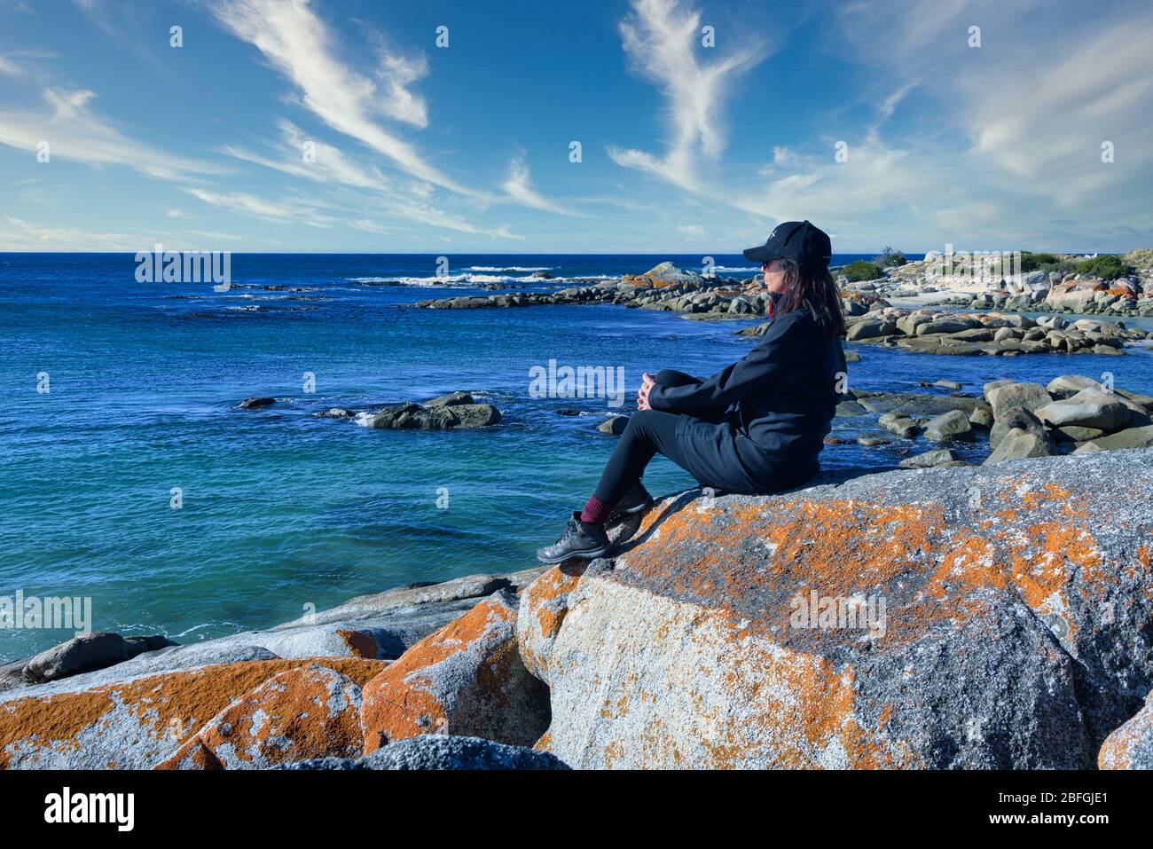 Female tourist sits atop a boulder covered with the characteristic orange lichens found in the Bay of Fires on Tasmania's rugged north-east coastline. Stock Photo