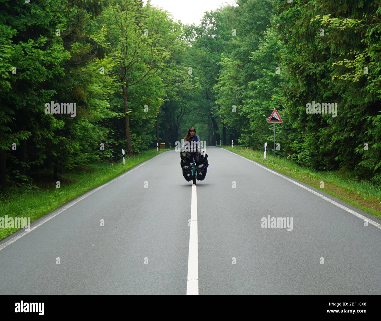 Bicycle world trip - steep mountain climb on a street in the forest. Woman on a bicyccle with full packed panniers. Stock Photo