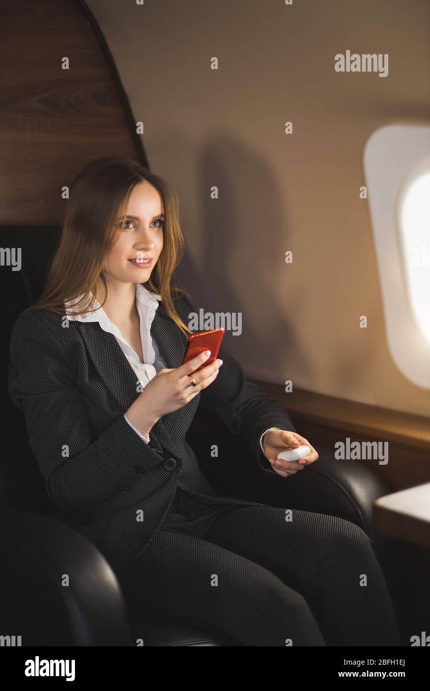 Beautiful young businesswoman traveling in private jet. Brown haired lady in white blouse, formal suit holding smartphone, sitting in comfortable seat Stock Photo