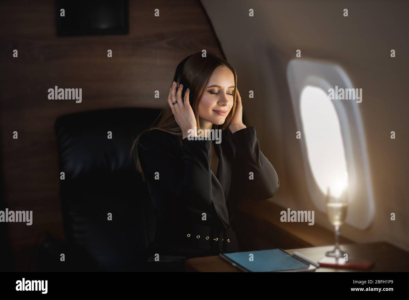 Happy caucasian young blonde woman in stylish black dress relaxing during flight in private jet, listening music using wireless headphones with closed Stock Photo