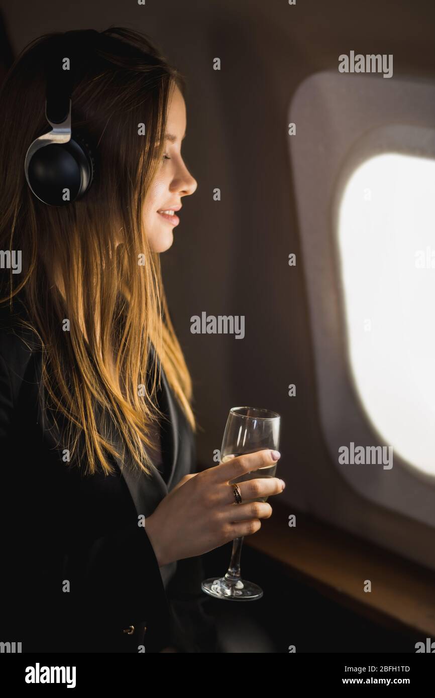 Vertical closeup shot of happy caucasian girl with long blonde hair, in black jacket dress, listening to audiobook with headphones, holding glass of c Stock Photo
