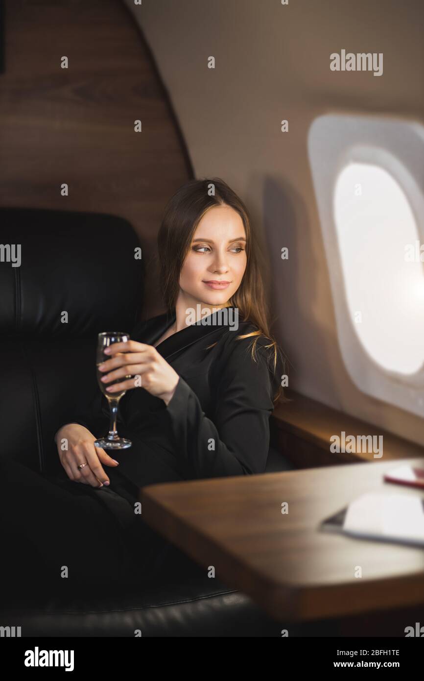 Beautiful happy young caucasian brunette girl flying in business class plane, sitting with glass of champagne in hand, smiling and looking at window. Stock Photo