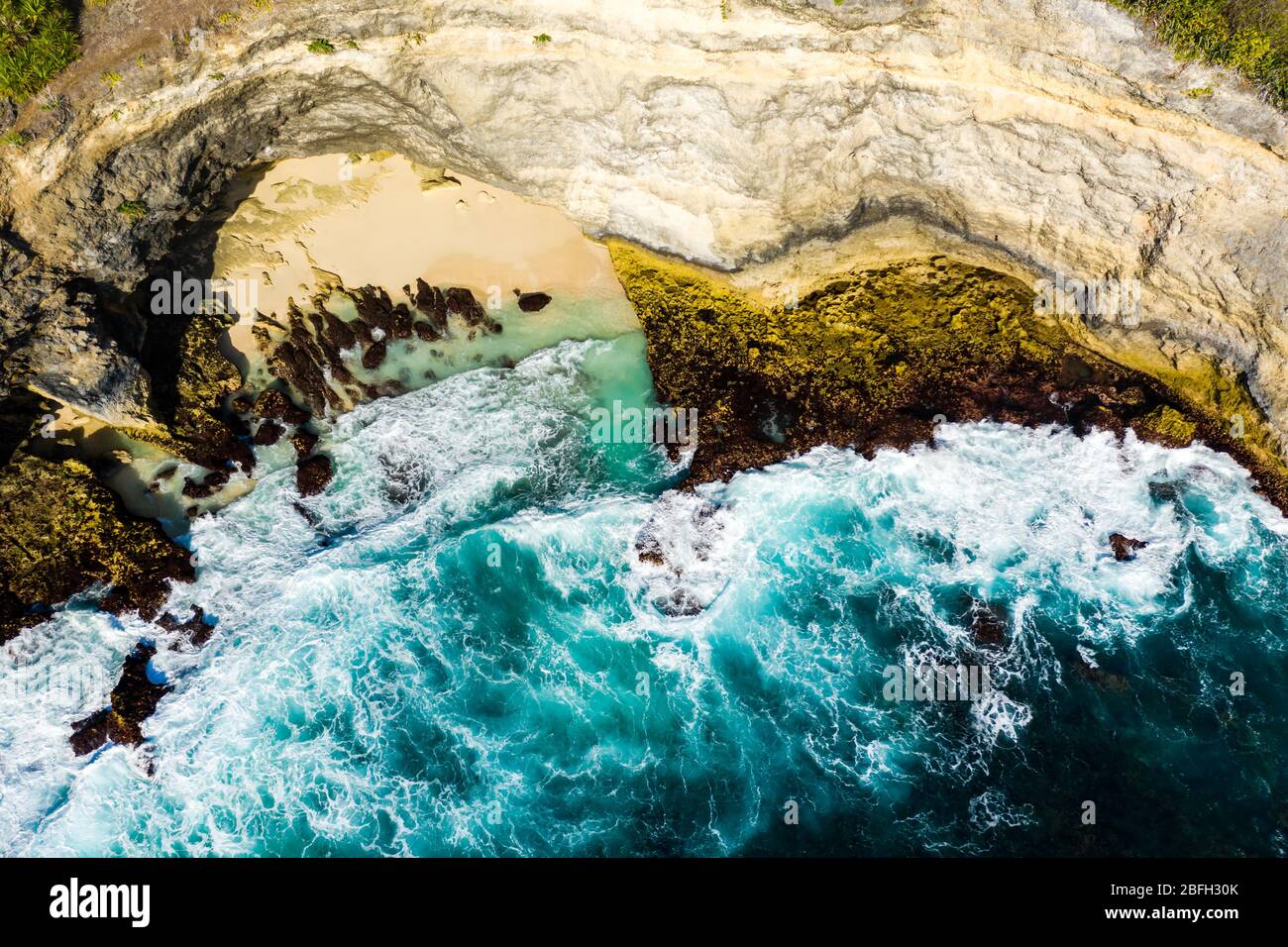 Top down aerial view of ocean waves crashing onto a small sandy beach surrounded by cliffs Stock Photo