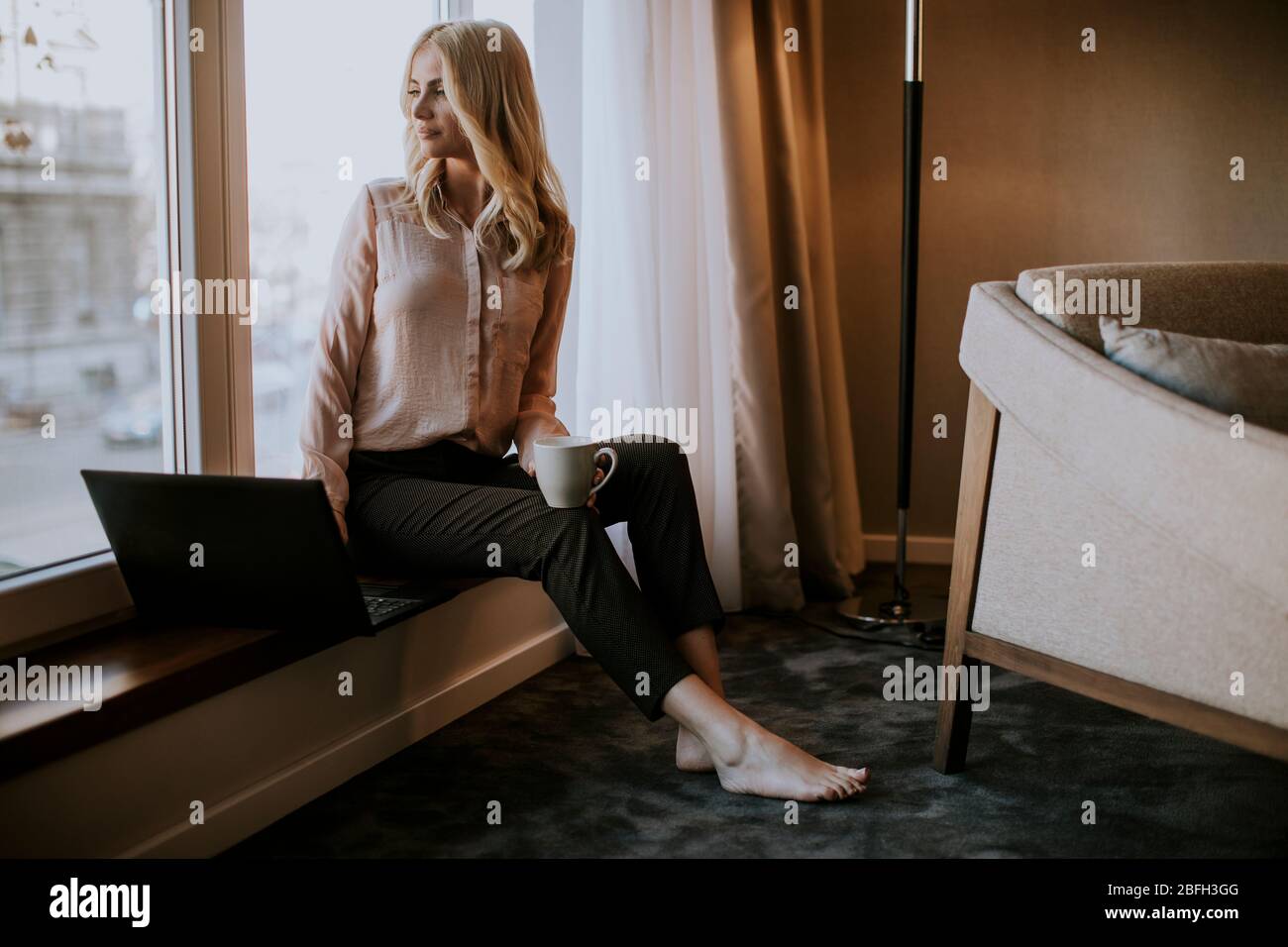 Pretty young woman working on laptop in living room on the window sill ...