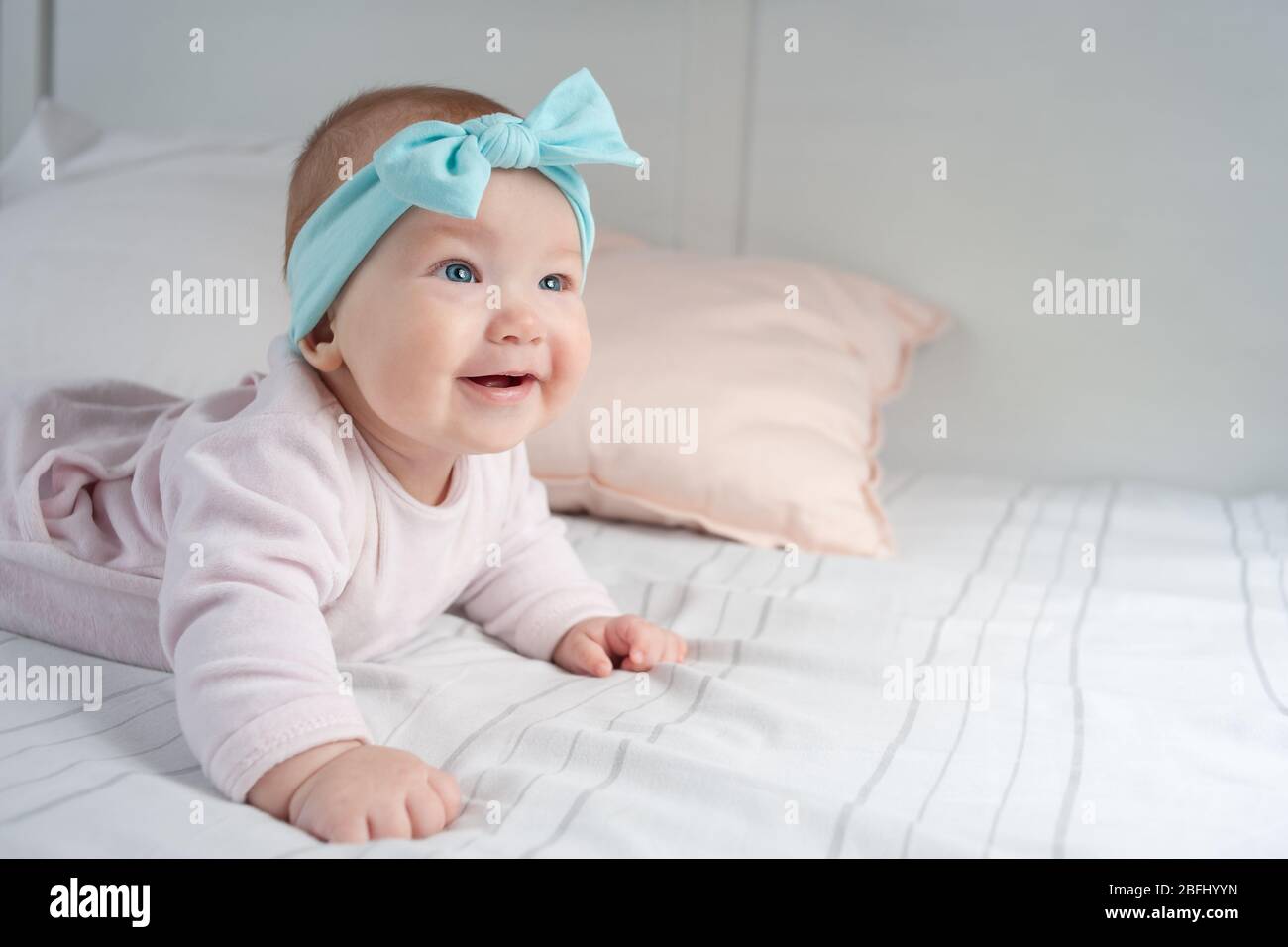 Newborn baby lies on its stomach and smiles. A child with blue eyes and a blue headband. Cute baby wearing lying on belly in nursery room. Beautiful s Stock Photo