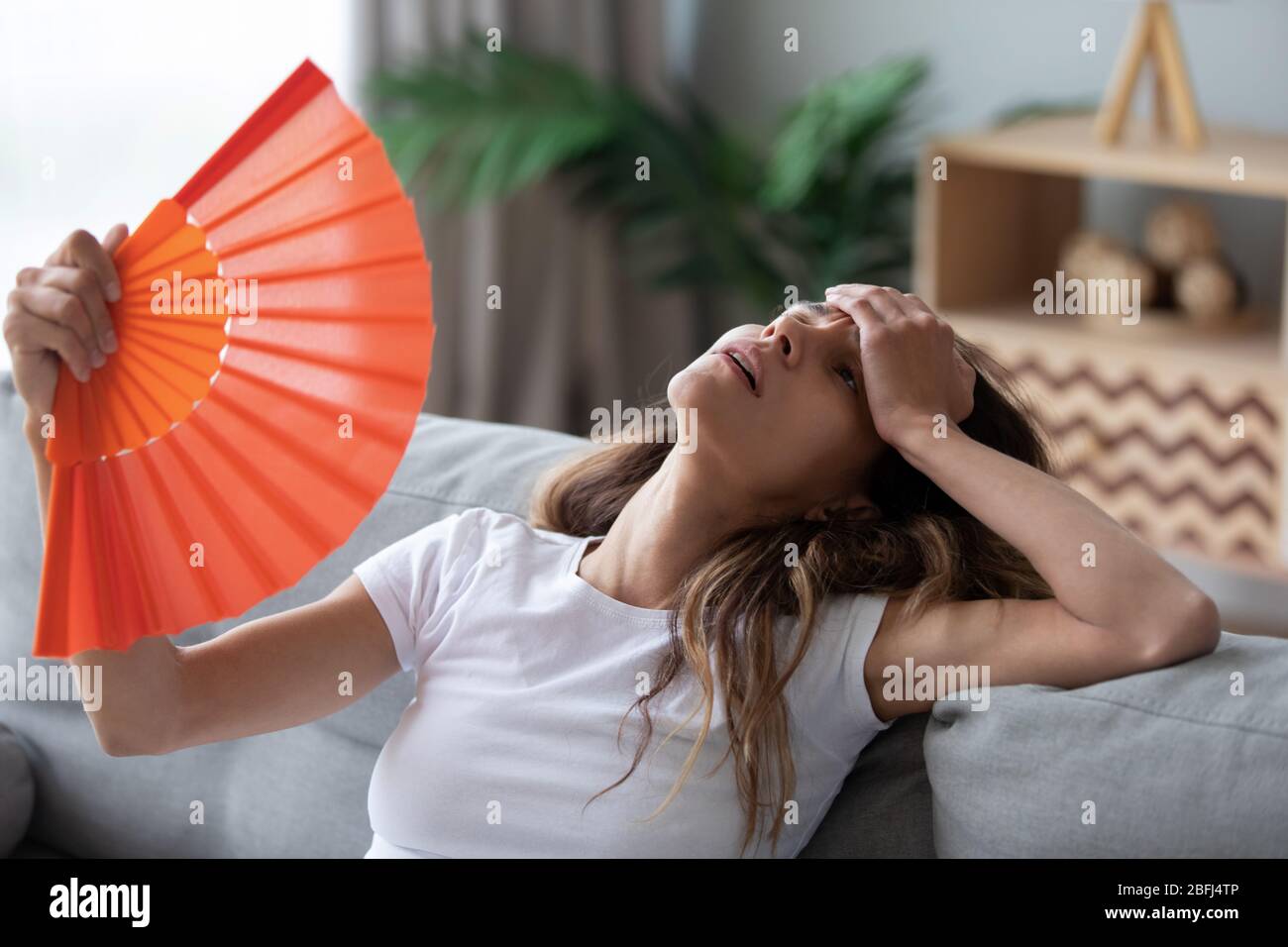 Overheated woman sitting on couch, waving fan, feeling unwell Stock Photo