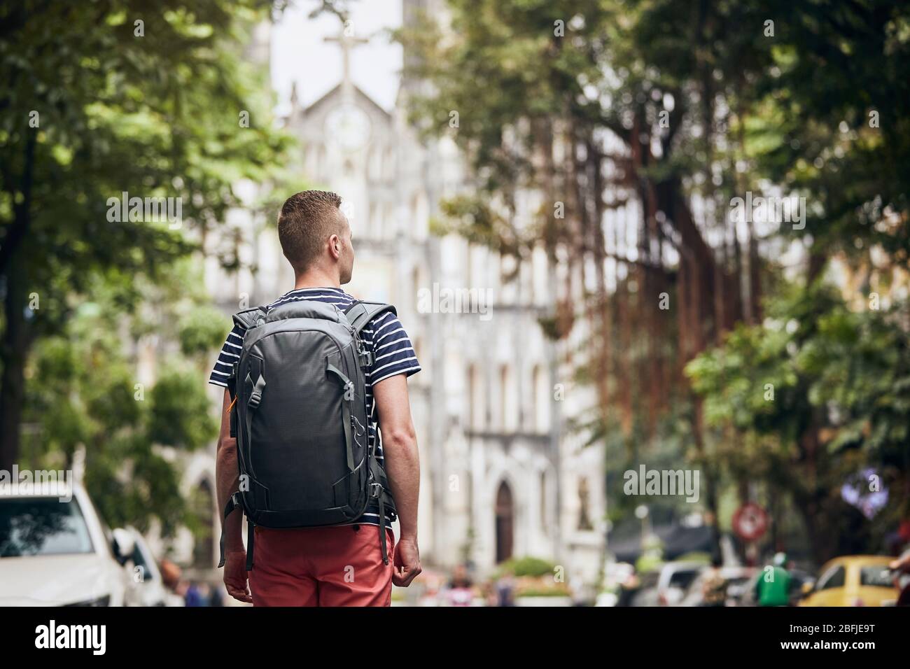 Traveler walking on street against cathedral. Rear view of young man with backpack in Hanoi, Vietnam. Stock Photo
