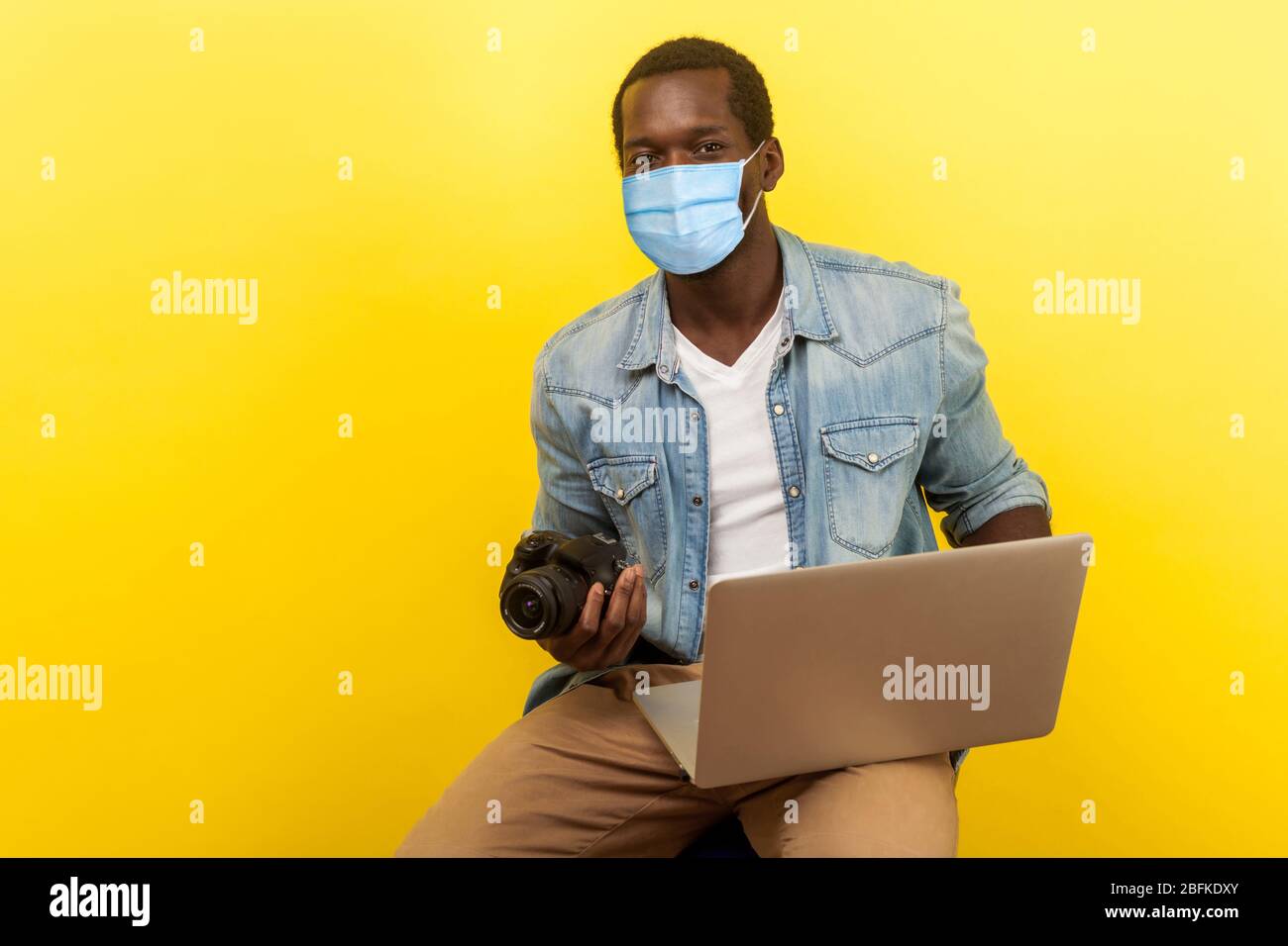 Portrait of energetic positive photographer with smile with surgical medical mask holding his camera and laptop, going to create video for blog. indoo Stock Photo