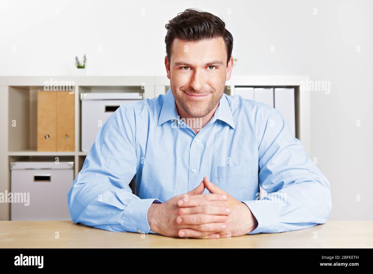 Attentive businessman sitting in office smiling at empty desk Stock Photo