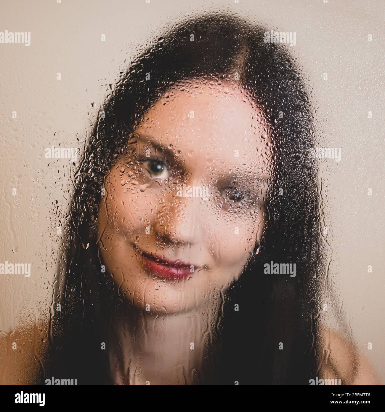 Face of happy girl with dark long hair and red lips behind a rain-covered glass window. Royalty free stock photo. Stock Photo