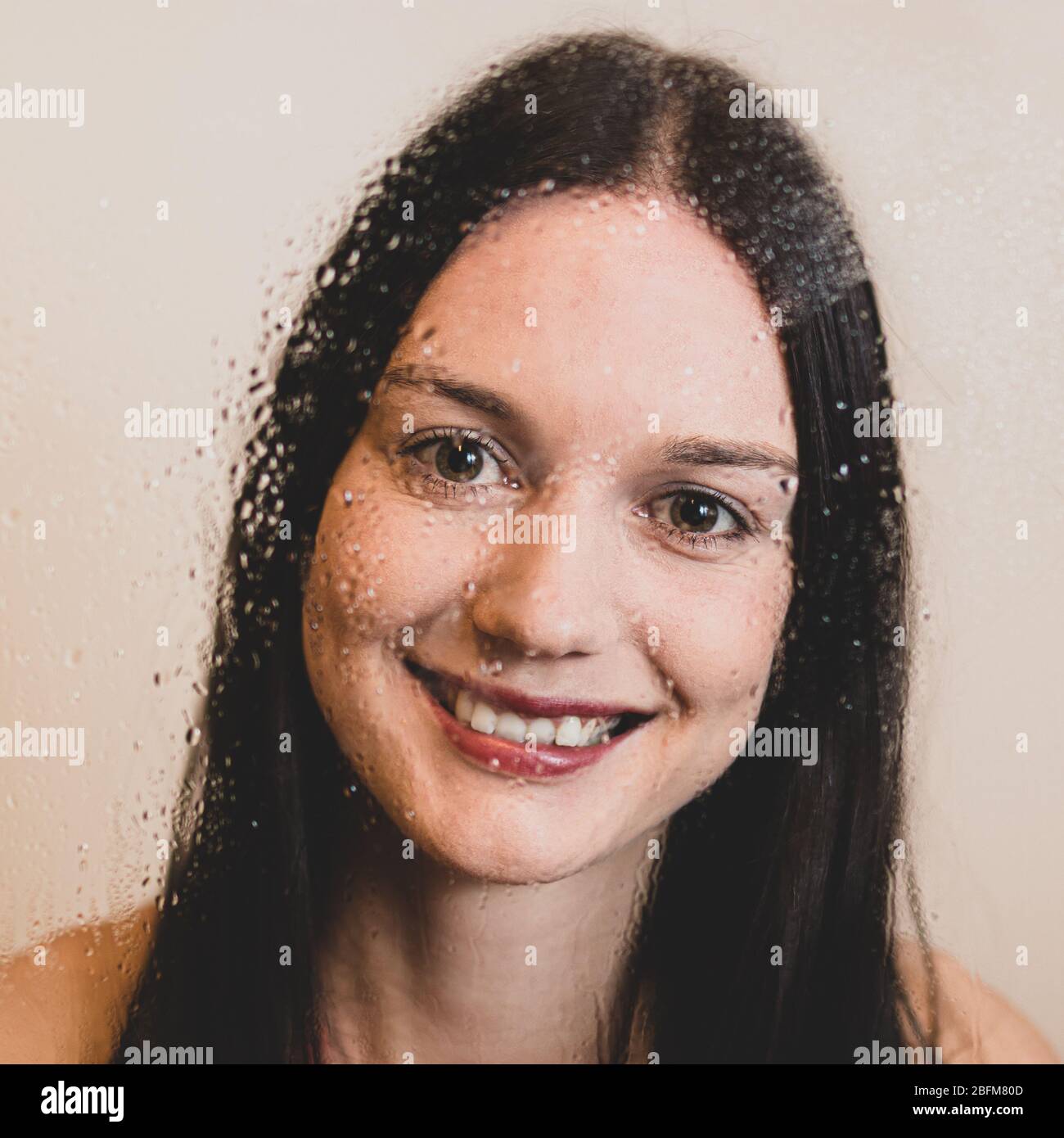 Face of happy girl with dark long hair and red lips smiling behind a wet glass window. Royalty free stock photo. Stock Photo