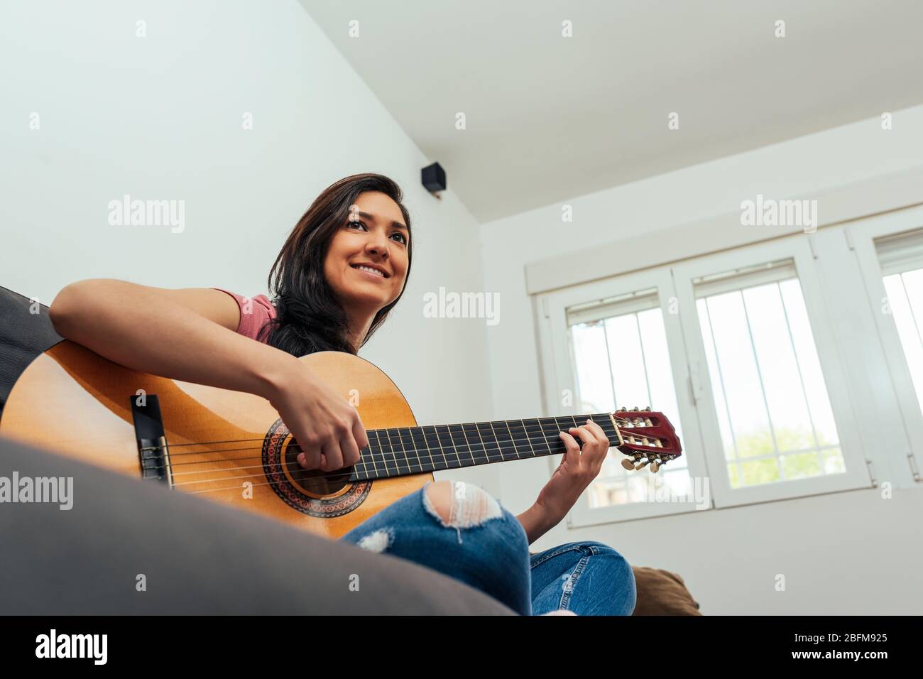 Latin woman playing guitar in the living room sitting on the couch. Stock Photo