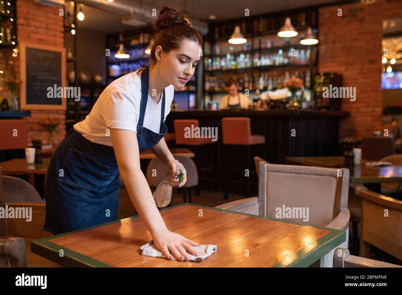 Pretty young waitress in apron bending over wooden table while using detergent and duster to clean it for new guests of restaurant Stock Photo