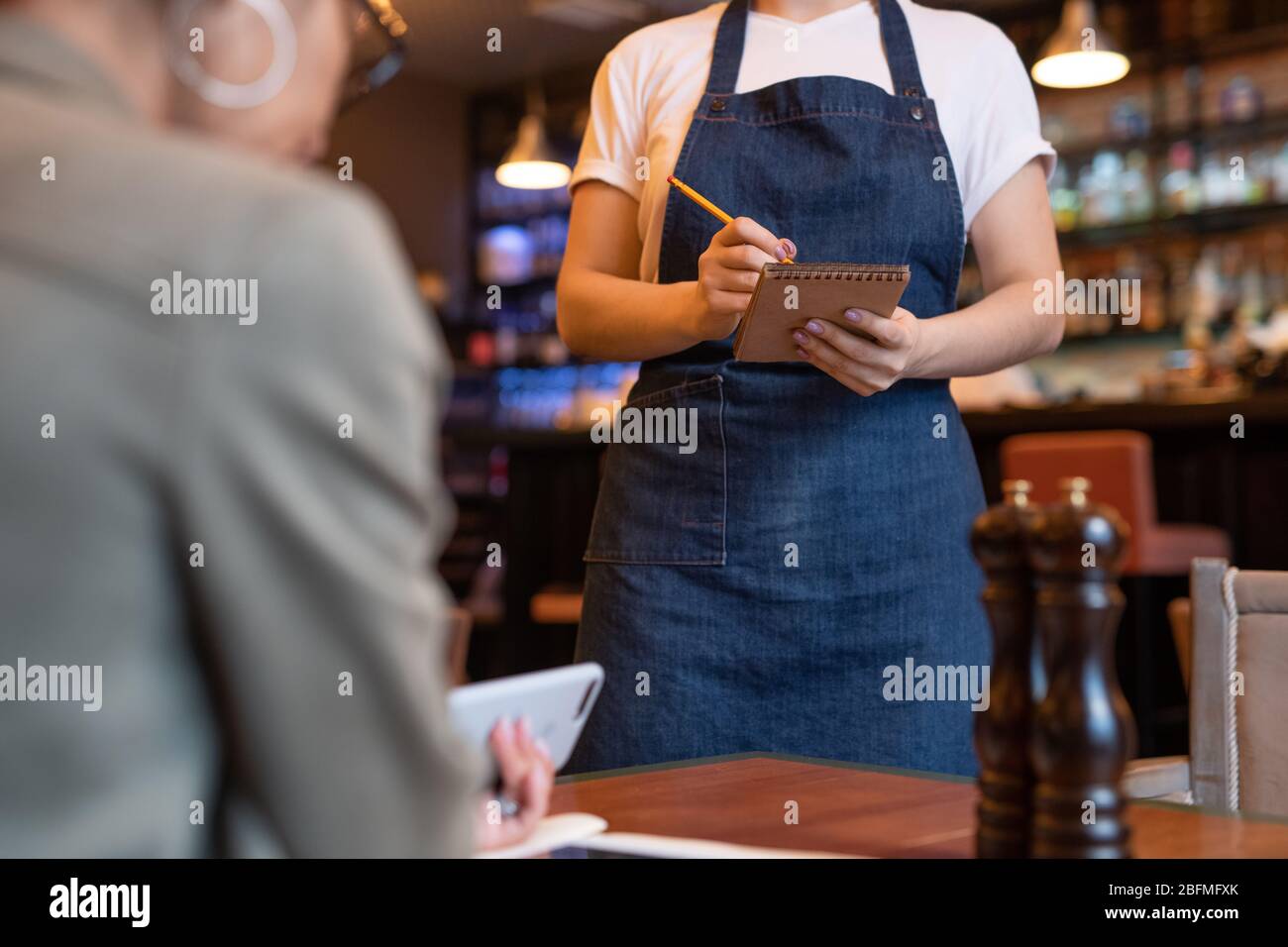 Young waitress in apron and white t-shirt writing don order of client in notepad while standing by her table in classy restaurant Stock Photo