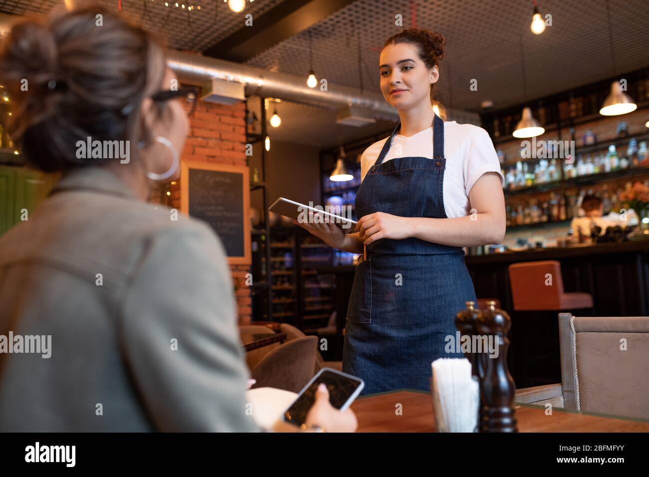 Young friendly waitress with touchpad standing by table with one of clients and taking her order on background of bar counter in restaurant Stock Photo