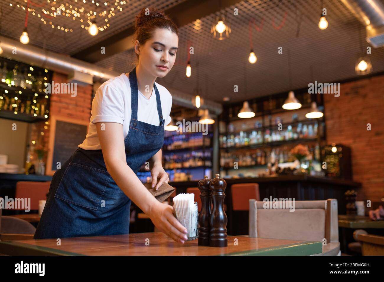 Young waitress of classy restaurant putting glass with bunch of toothpicks, salt and pepper on one of tables while preparing it for guests Stock Photo
