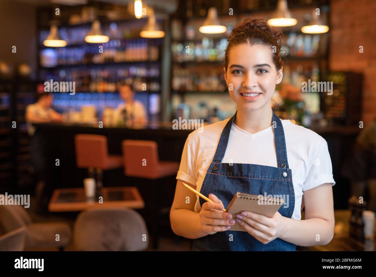 Young smiling waitress in workwear standing in front of camera in luxurious restaurant and going to writie down order of client Stock Photo