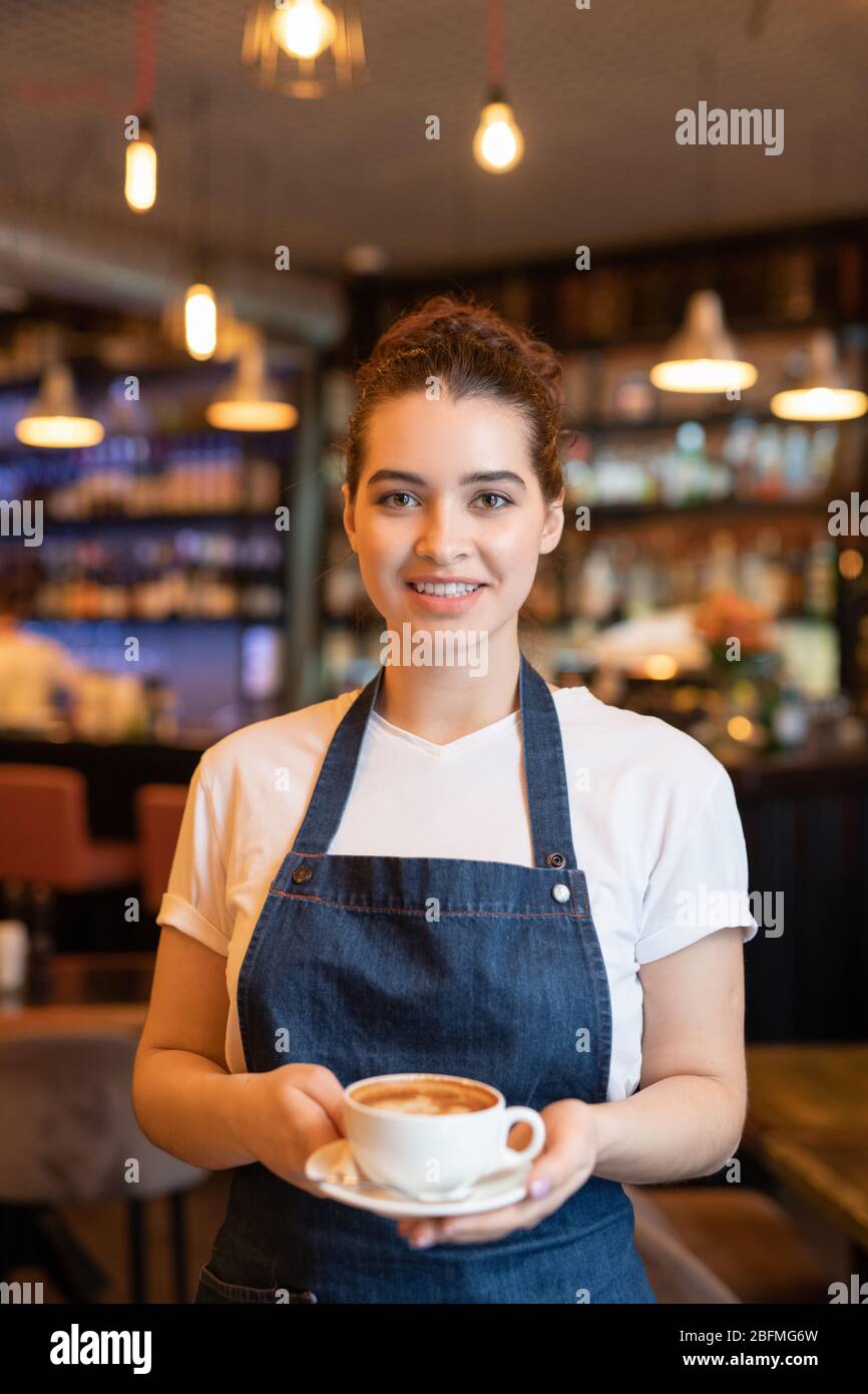 Young cheerful brunette waitress in white t-shirt and dark blue apron passing cup of fresh cappuccino while standing in front of camera Stock Photo