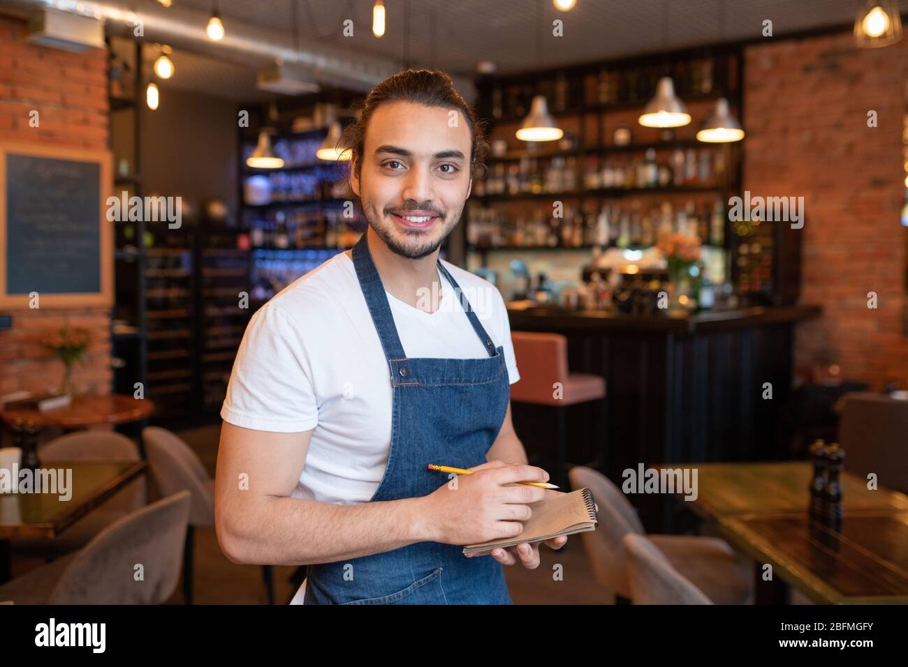 Happy young waiter in white t-shirt and dark blue apron looking at you with toothy smile and writing down your order in restaurant Stock Photo