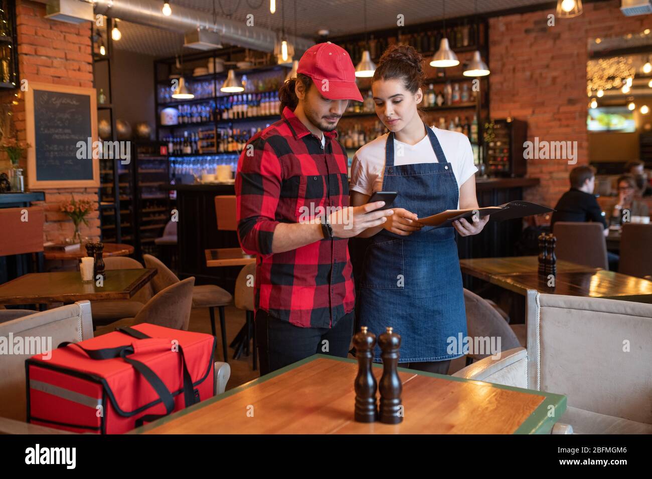 Young courier with smartphone and waitress with menu standing by table and checking online orders before delivering them to clients Stock Photo