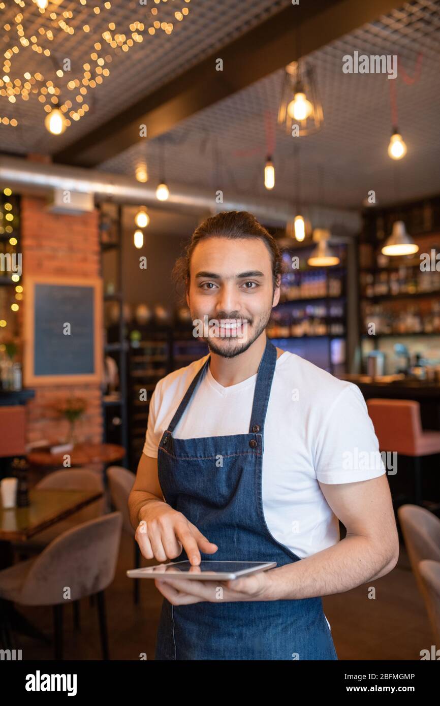 Young cheerful and friendly waiter of classy restaurant scrolling through online orders in digital tablet while waiting for new guests Stock Photo
