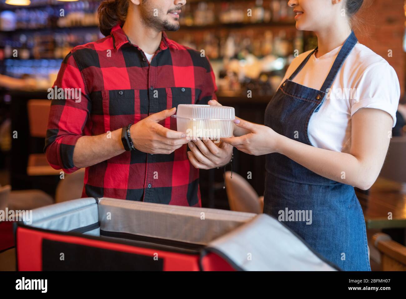 Young courier in red flannel taking container with resh food ordered by one of clients from hands of waitress helping him with packing Stock Photo